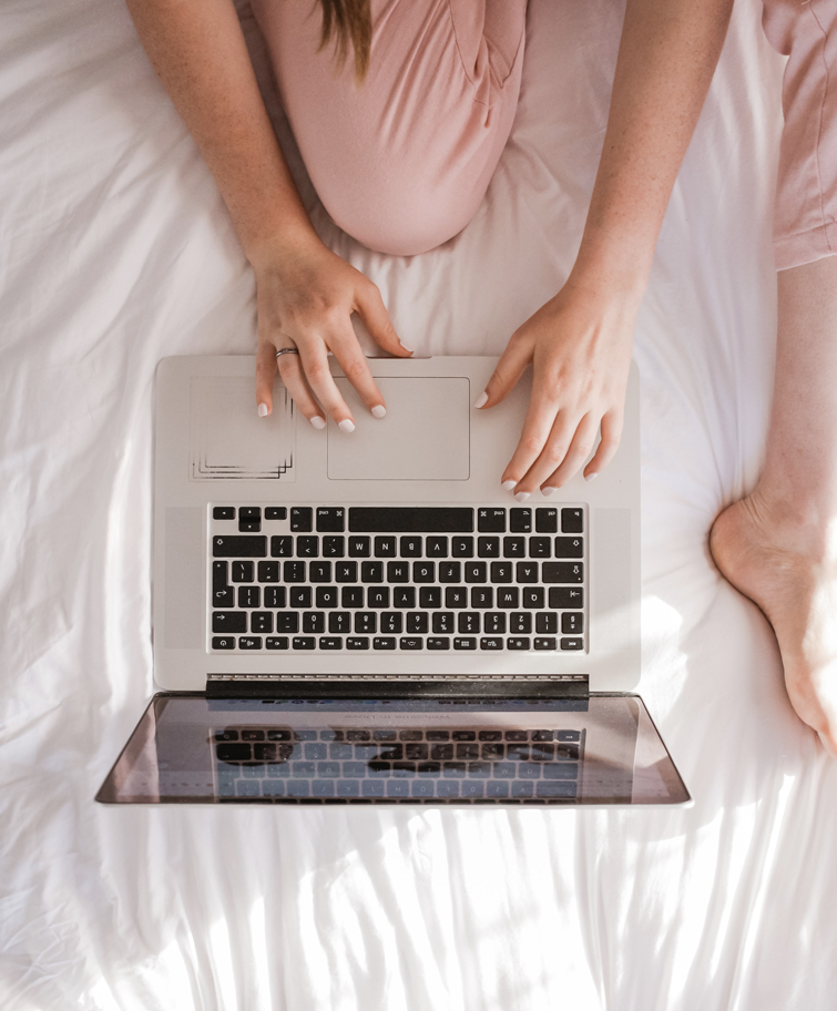 Frau mit rosa Hose sitzt mit ihrem Computer auf ihrem Bett, die gerade online shoppen macht