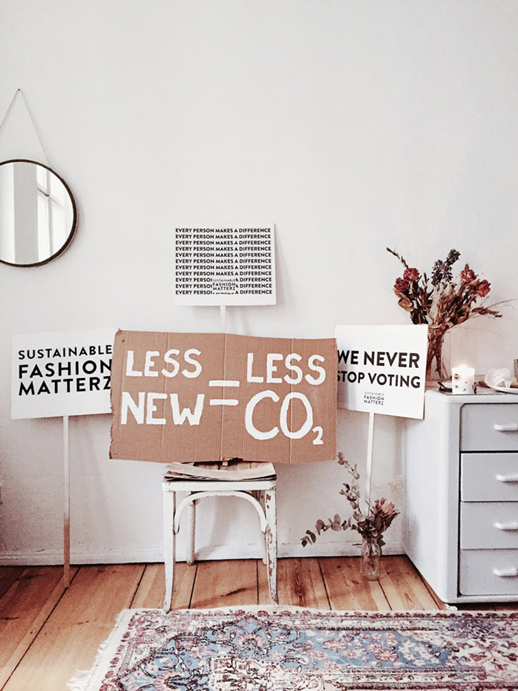 Zimmer mit einem Spiegel, einem Blumenstrauß in einer Vase und Schild Ûber Schnelle Mode auf einem Holzstuhl mit der Aufschrift "LESS NEW = LESS CO2"